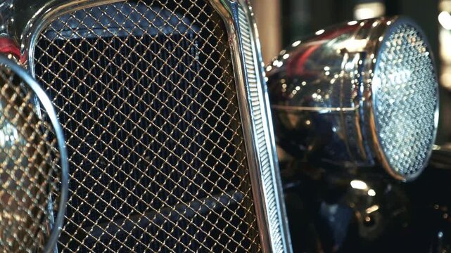 A close-up view of the front section of a vintage vehicle showing a shiny chrome radiator grill and a large circular headlight on polished black bodywork.