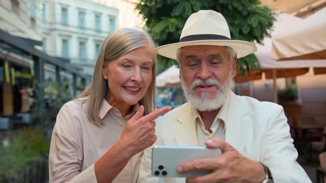 Happy Caucasian elderly couple grandparents blogging with mobile phone on city street retired family woman man talking call live stream smartphone online vlog traveling on holidays near cafe outdoors