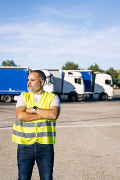 Male truck driver wearing a safety vest and jeans, standing with arms crossed at a truck stop with vehicles in background