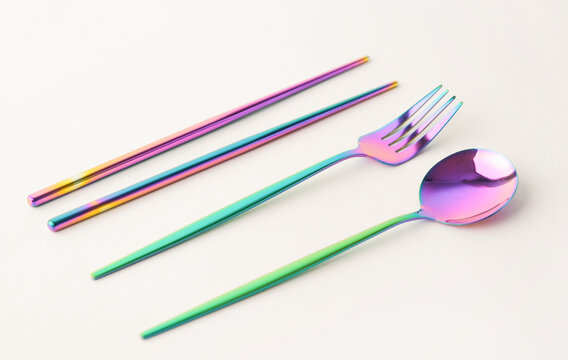 iridescent metallic sticks , fork and spoon resting on a glossy white surface, vibrant rainbow gradient reflecting soft pink and blue light