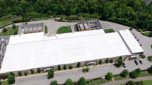 A long white roofed data center with rooftop fan arrays, ground level cooling units and a fenced generator yard, transmission lines running past the treeline. Aerial lateral wide shot