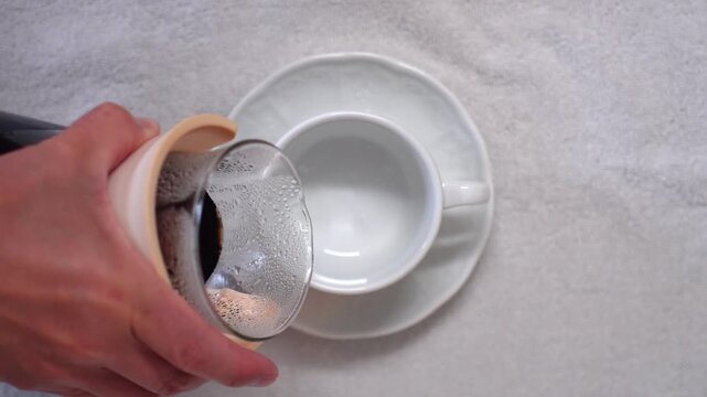 Top view of freshly brewed black coffee poured from a glass server into a white cup on a saucer. Hand pouring drip coffee, cafe and breakfast scene, empty cup to full cup.