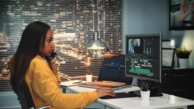 A woman with long dark hair talks on smartphone while working on video editing software at a white desk in a modern urban office with city lights visible through large window at night.