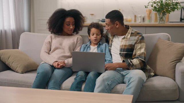 Happy African American family parents talking little boy child using notebook laptop computer indoors living room explaining software internet app technology mom dad son home digital parental control