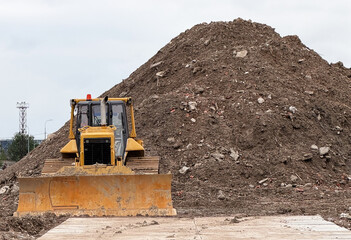 Heavy industrial yellow crawler bulldozer working at a construction site. Powerful earthmoving machinery vehicle clearing ground near a huge pile of soil dirt and stones under an overcast sky.  © Dmitry Presnyakov