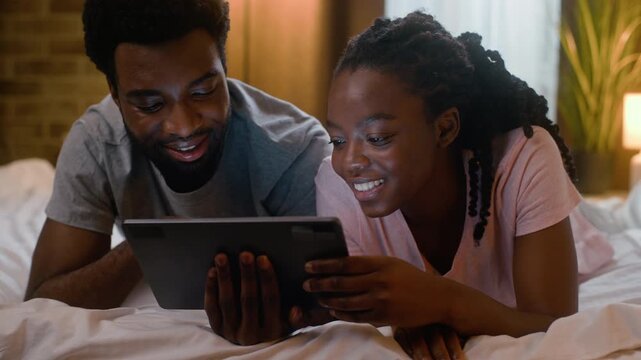 Happy African American couple lying bed watching movie digital tablet together laughing and cuddling at home bedroom late evening romantic family relationship woman girlfriend man boyfriend having fun