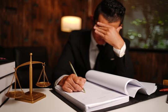 Stressed Lawyer Covering Face while Writing Legal Documents with Scales of Justice