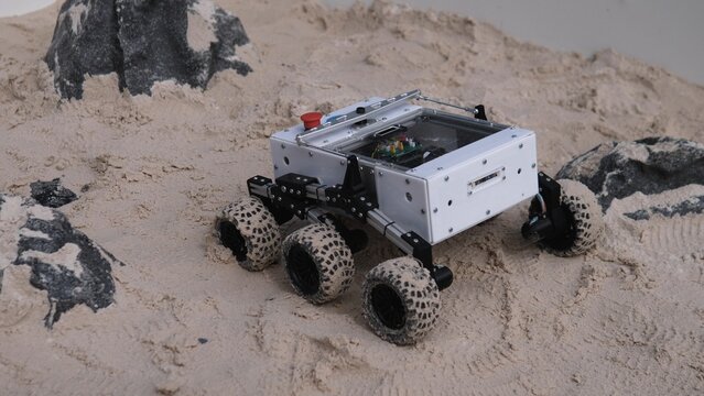 Compact wheeled robot moves over fine sand near dark rocks, showing metal chassis, clear top cover, exposed circuitry, and dust coated tires in controlled indoor testing conditions