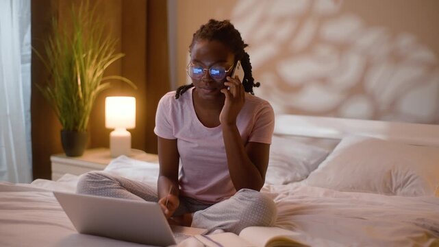 Multitasking African American business woman talking smartphone call and laptop at home bedroom night female freelancer speaking mobile phone typing computer writing notes working online communication