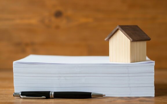 Miniature wooden house model on a stack of white paper with a black pen in front, on a wooden surface