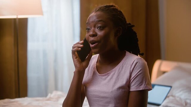 Laughing African American woman talking on mobile phone call sitting in bedroom at home cheerful ethnic female girl smartphone conversation chatting friends gossiping happy smile indoor late evening
