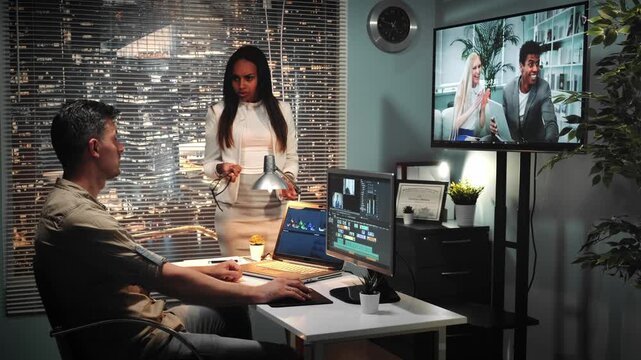 Three professionals collaborate in a modern office with a large vertical screen showing remote colleagues engaged in discussion. Indoor daytime setting.