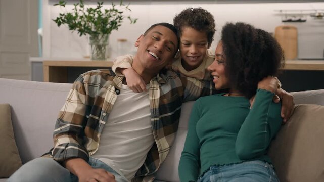 Happy African American family mother father and little boy child smiling embracing together loving parents mom dad kid con laughing talking relaxing weekend on sofa couch indoors home living room