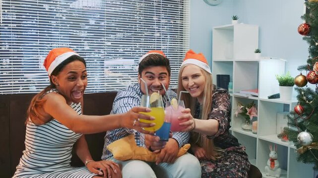Three adults wearing Santa hats smile and hold colorful cocktails while seated on a sofa in a decorated living room with a Christmas tree and holiday decor.