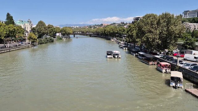 Title: Tourist Boats Navigating Kura River Near Bridge of Peace and Rike Park in Tbilisi