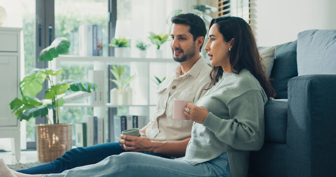 Happy young adult Hispanic couple drinking coffee and relaxing sit on floor in living room at home. Domestic lifestyle, positive weekend leisure, cheerful relationship bonding, family love concept.