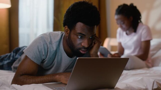 African American couple working laptop and digital tablet at night in bedroom husband wife typing computer device online gadget addiction shopping reading news evening ethnic married family at home