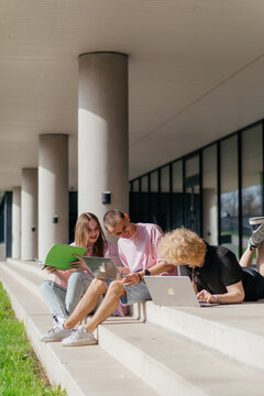 Group of students studying and using laptops outside a modern university building on a sunny day
