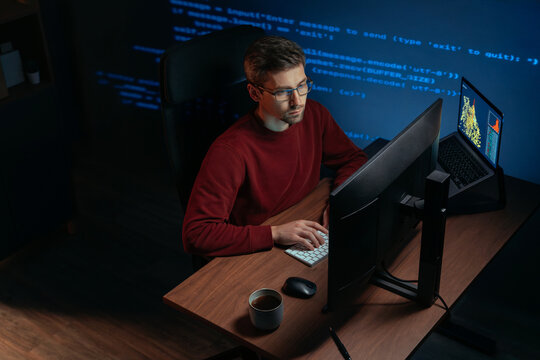 Software engineer wearing eyeglasses working at multi-screen desk in a dark office, focused on code and graphs. Programmer works late at night on a demanding data science and AI task. Copy space