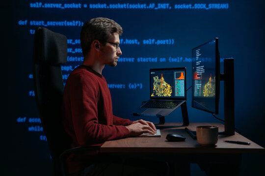 Side view, coder at a dark office desk works across multiple screens at night, with programming script on a background. Data scientist working on a machine learning or artificial intelligence task.
