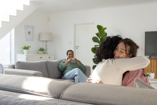 African female family hugging in living room while senior sitting on gray sofa holding white mug