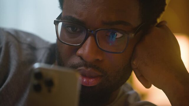 Stunned African American man in eyeglasses using smartphone lying in bed looking mobile phone screen reading news browsing internet close up portrait male guy user client customer home bedroom evening