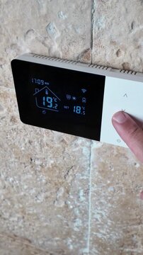 Vertical view of a hand lowering the temperature on a digital heating boiler controller to reduce home energy consumption, illustrating smart domestic climate control, conservation, and efficiency.