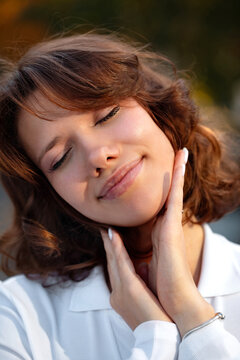 Young woman with closed eyes gently holding her neck outdoors in a white blouse for a peaceful lifestyle portrait