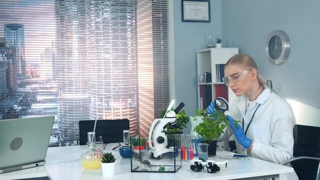 A female scientist wearing safety glasses and gloves examines a green plant leaf with a magnifying glass at a white laboratory table filled with scientific instruments during daytime.