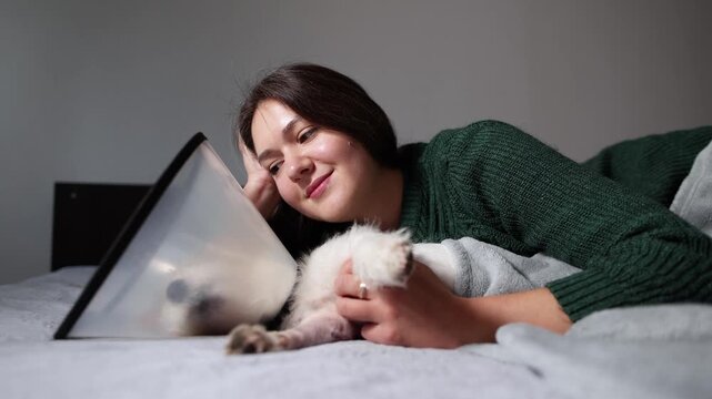 Low angle shows woman embracing Jack Russell after leg surgery. She gently strokes dog on bed while cone collar and calm care frame recovery.
