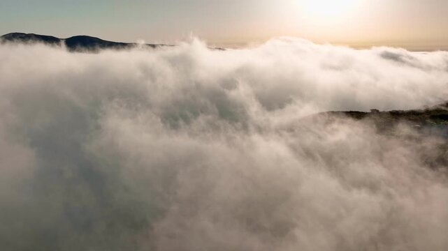 A drone flies above the clouds, showcasing a cloud inversion over Fanal forest in Madeira, Portugal during sunrise. The landscape reveals hills and dense trees.