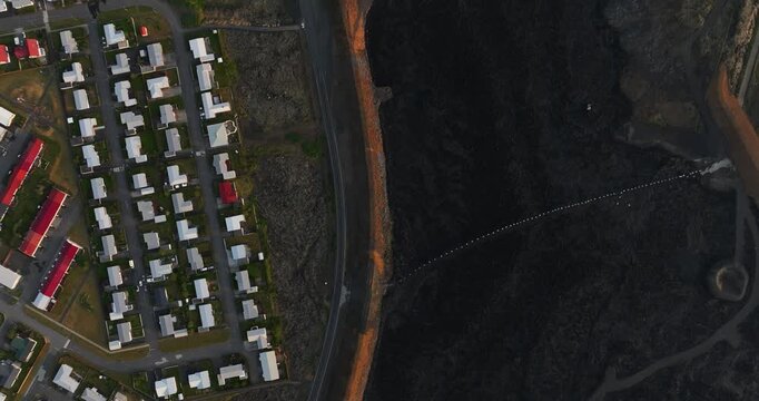Top-down drone view over Grindavik, Iceland, showing a dense housing grid beside a long orange defense berm and vast black lava field crossed by service tracks.