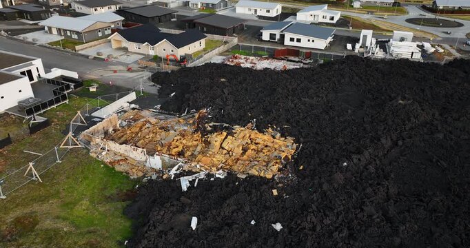 Oblique drone view in Grindavik, Iceland, showing a home reduced to exposed timber and rubble as hardened black lava surrounds the property beside a green lawn and nearby houses.
