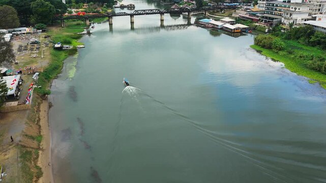 The steel bridge over the River Kwai carries its curved spans across the water at Kanchanaburi, Thailand, a long tail boat trailing a wake past floating restaurants. Aerial drone wide shot