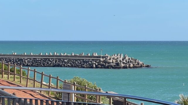 A stone and concrete breakwater lined with dolos interlocking wave-breaking structures extends into a turquoise ocean under a clear blue sky.