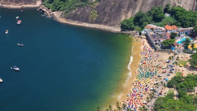 View from Sugarloaf Mountain over Vermelha Beach, its sandy shore dotted with colorful umbrellas and tiny figures of people in the deep blue waters of the Atlantic ocean, seen from high above