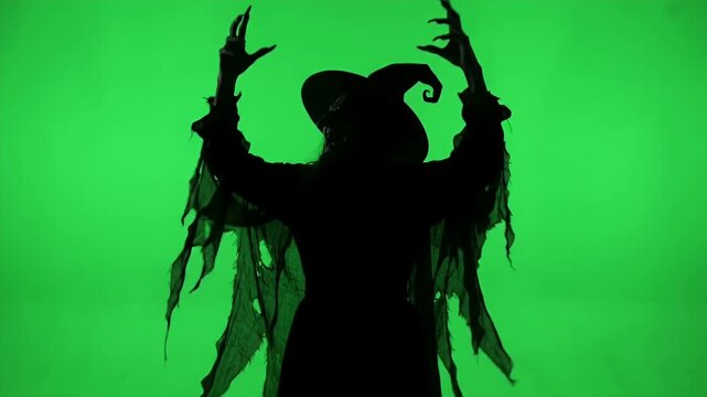 Silhouette of a Wicked Witch Cackling on Green Screen - A silhouetted witch in a pointed hat with ragged robes and long claws raises her hands as if cackling with wicked glee.