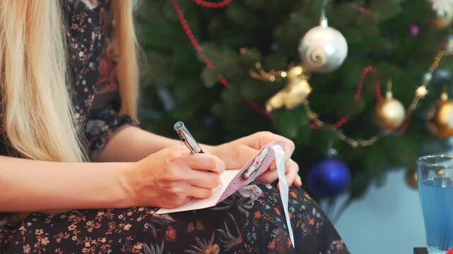 A woman with long blonde hair writes a postcard while seated indoors near a decorated Christmas tree with ornaments and glasses on a table.