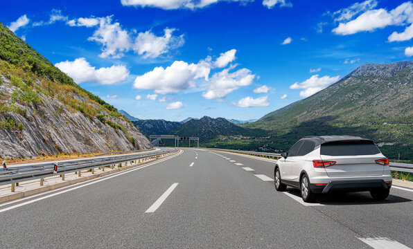 A modern white car drives along a scenic highway winding through picturesque mountain landscapes under a bright blue sky.