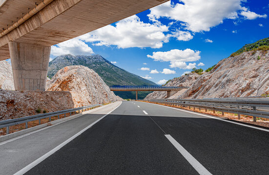A wide asphalt highway passes under a concrete bridge through a scenic rocky mountain landscape under a bright blue sky with white clouds.