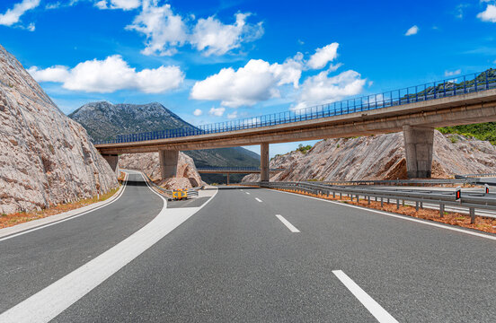 An empty modern highway passes under a concrete overpass through a scenic mountain landscape under a bright blue sky with white clouds.