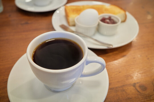 A white cup of black coffee rests on a wooden table with blurred buttered toast a boiled egg and sweet red bean paste in the background Ideal for food blogs and cafe menu marketing