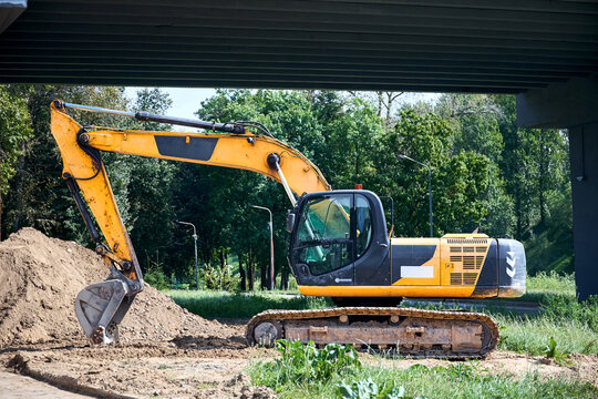 Yellow tracked excavator parks on dirt ground under concrete bridge structure. Heavy construction machinery with bucket stands near embankment on industrial building site.