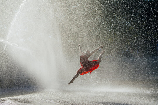 White dancer leaping through fountain spray in red dress outdoors