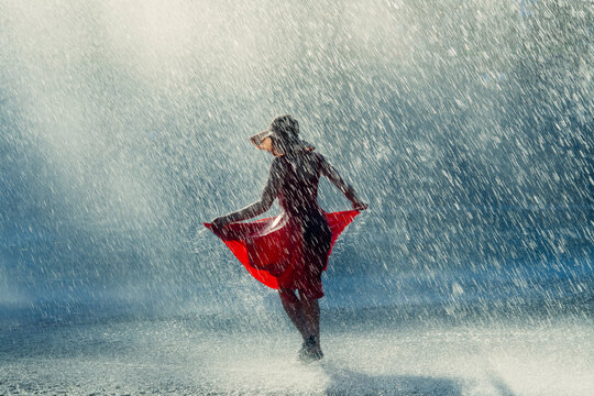 White woman dancing joyfully in rain wearing red dress and sun hat