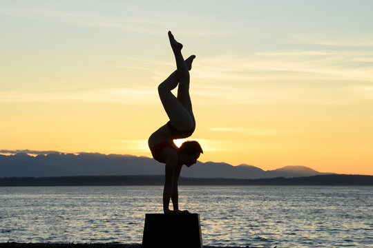 White woman doing handstand on box at ocean sunset with mountains