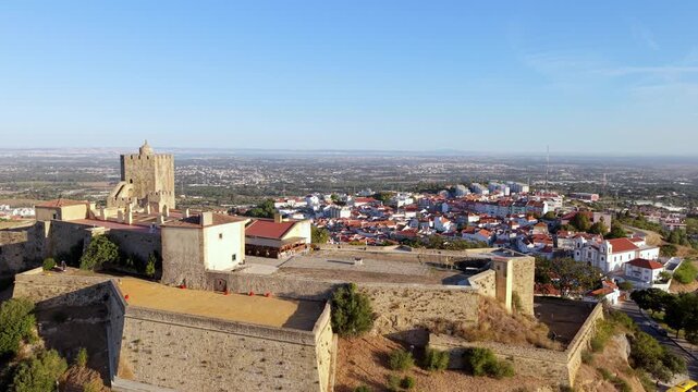 Aerial view of historic castle overlooking colorful town. Ancient stone walls frame scenic landscape. Town spreads below with red-roofed homes. Bright blue sky enhances warm tones