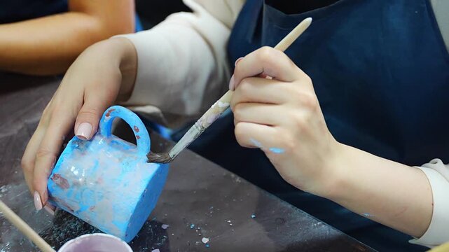 Woman applying blue glaze to handmade ceramic mug with brush. Pottery glazing process represents ceramic craft and handmade production