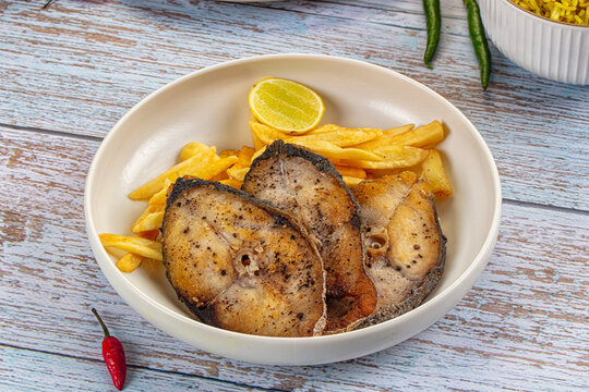 Grilled kingfish steak served with golden french fries and lemon wedge on rustic blue wooden table, red chili pepper garnish