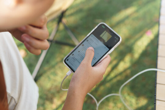 A woman uses a smartphone to browse the internet while it's still plugged in as a charger. This is a potentially dangerous use of the device. The photos depict daily life in the backyard.Spare battery
